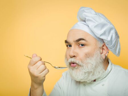 Bearded man cook in hat tasting food with spoon in studio on yellow backgroundの写真素材