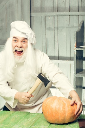 Bearded man cook in chef hat with axe and pumpkin on wooden backgroundの写真素材
