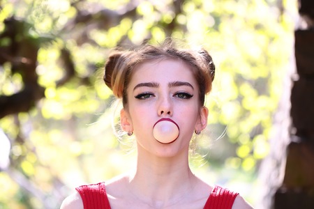 young pretty woman with chewing gum in the garden with fresh spring leaves on trees on blurred...