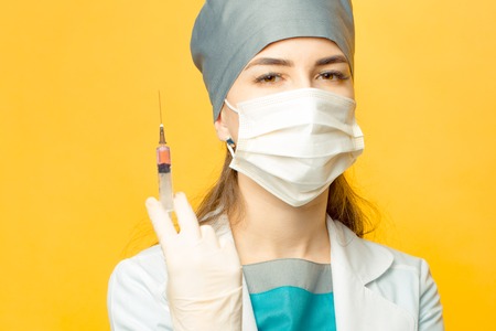 Beautiful young doctor in mask and medical uniform holding syringe with liquid on yellow backgroundの写真素材