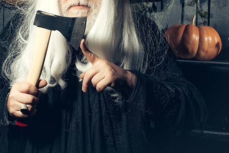 Bearded senior man in long white wig with axe on wooden background with pumpkinの写真素材