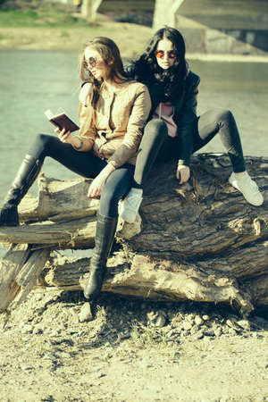 Young girls in stylish sunglasses with beautiful long hair reading book sunny day outdoor sitting on big wooden root near waterの写真素材