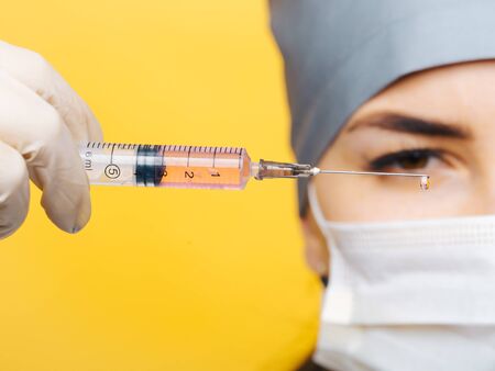 Young doctor in mask holding syringe with liquid closeup on yellow background