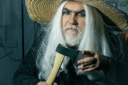 Bearded senior man in long white wig and straw round hat with axe on wooden backgroundの写真素材