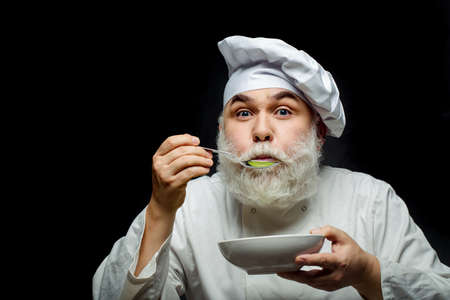 Bearded man cook in hat tasting food with spoon in studio on black background, copy spaceの写真素材