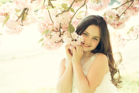Beautiful small girl in white glamour dress with long hair and smiling face in spring pink flower blossom sunny dayの写真素材