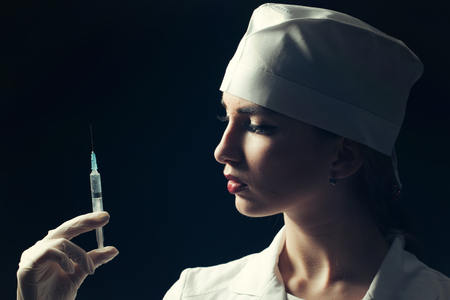Young doctor with beautiful face in medical uniform holding syringe with liquid on black backgroundの写真素材