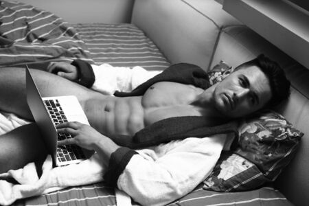 Young man with muscular body lying on bed near laptop at the bedtimeの写真素材