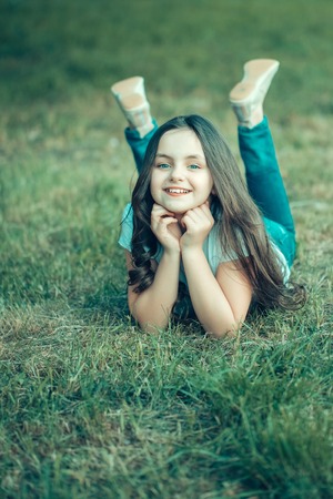 Beautiful small girl with smiling face and long hair lying on green grass in spring sunny dayの写真素材