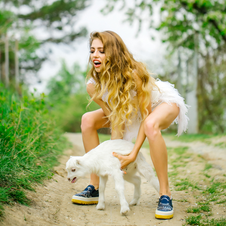 Young woman with beautiful face and long curly hair in glamour dress holding cute white small goat in country sideの写真素材