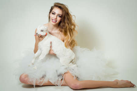 Young smiling woman with sensual face and long curly hair topless in glamour skirt holding cute white small goat in studioの写真素材