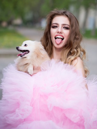 Young woman with beautiful face and long curly hair in glamour pink dress showing tongue holding cute small dog outdoorの写真素材