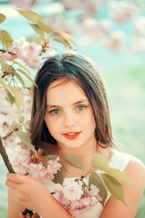 Beautiful small girl in white glamour dress with long hair and smiling face in spring pink flower blossom sunny dayの写真素材