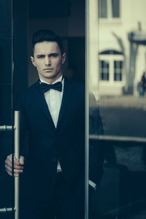 Young handsome man in elegant suit with bow tie stands at glass entrance doorの写真素材