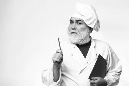 Bearded senior cook man pointing recipe at the cookbook at the kitchen in hat, black and whiteの写真素材