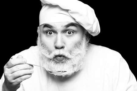 Bearded man cook in hat tasting food with spoon in studio, black and white