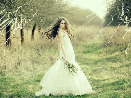 Beautiful young bride in white wedding dress with flowers in field with ribbons on trees outdoorの写真素材
