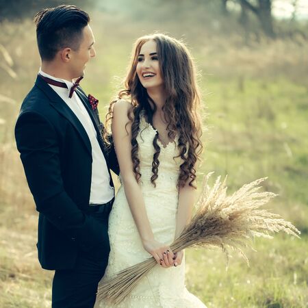 Happy newlywed couple posing and smiling in wedding dress and suit with flowers in field outdoorの写真素材