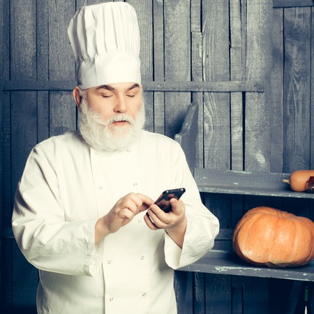 Bearded man cook with mobile phone in chef hat and uniform  in studio on wooden background with pumpkinの写真素材