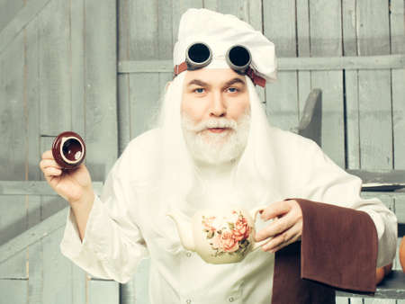 Bearded man cook in chef hat and glasses holding teapot and napkin on wooden backgroundの写真素材