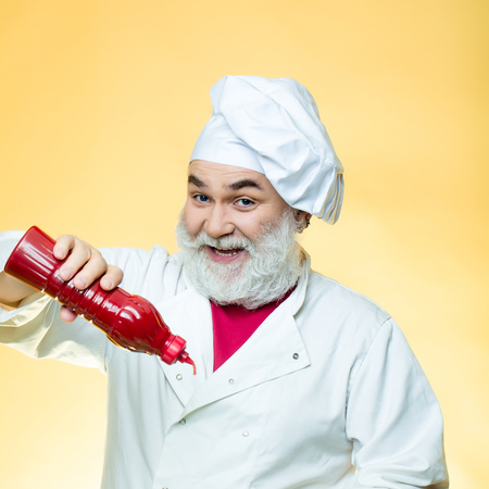 Bearded chef with ketchup bottle and smiling face in white uniform and hat on yellow background