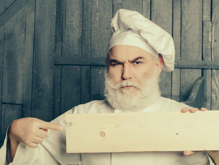 Bearded man cook with serious face holding and showing on empty wooden plate in chef uniformの写真素材