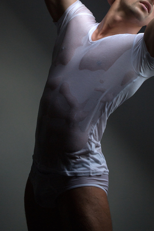 Sexy young man with muscular body in wet shirt posing in trunks near windowの写真素材