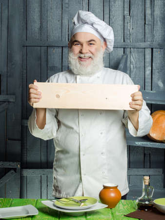 Bearded man cook with smiling face in studio holding empty wooden plate preparing food with sauce in chef uniformの写真素材