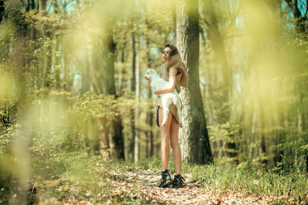 Young woman with beautiful face and straight body in beige burlap short dress with hood holding cute white small goat in green forestの写真素材