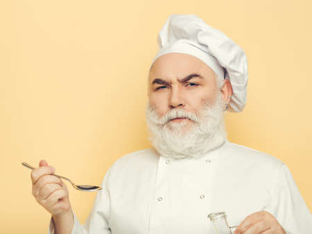 Bearded man cook in hat tasting food with spoon in studio on yellow background, copy spaceの写真素材