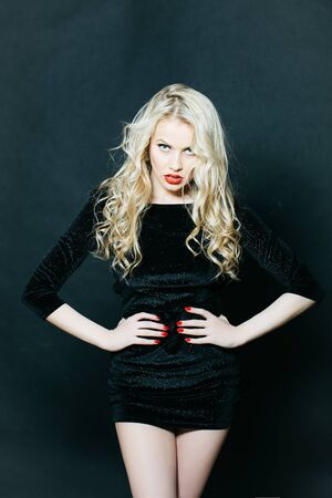 Attractive sexy woman with blonde curly hair posing in black elegant dress in studio on grey backgroundの写真素材