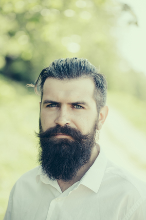 Handsome young stylish hipster man with long beard in white shirt standing outdoor on natural background sunny day outdoorの写真素材