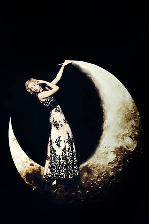 Beautiful young woman with blonde hair and red lips posing on crescent moon in long evening elegant dress with lace on black backgroundの写真素材