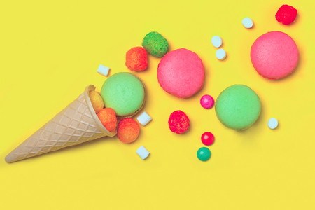 Colorful macaron pink and green color drop out of ice cream cone of waffle with many small jelly candies and dragee with marshmallow on yellow backgroundの写真素材
