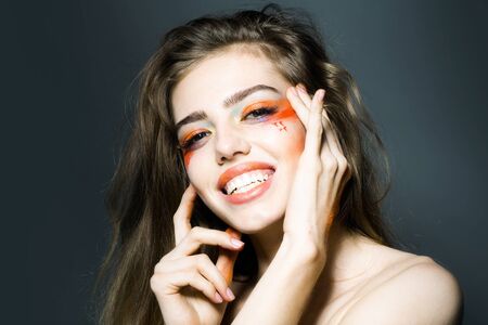 Young girl with hands near pretty smiling face and bright orange makeup in studio closeup