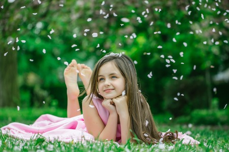 Beautiful little girl in pink dress with long brunette hair and smiling face lying barefoot on green grass in spring flower blossom petals outdoorの写真素材