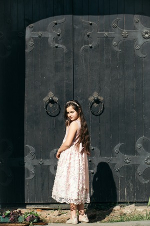 Small girl in pink dress and wreath standing near old wooden door with iron details sunny day outdoorの写真素材