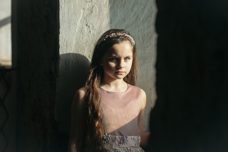 Pretty little girl in dress and wreath with long brunette hair and pensive face standing near building wall grey color in sun lightの写真素材