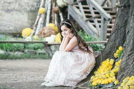 Pretty little girl in dress and wreath with long brunette hair and thoughtful face sitting near tree with floral decoration of yellow roses outdoorの写真素材