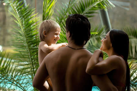 Happy family couple of young smiling woman and muscular man holding son on hands near swimming pool with green palm leavesの写真素材