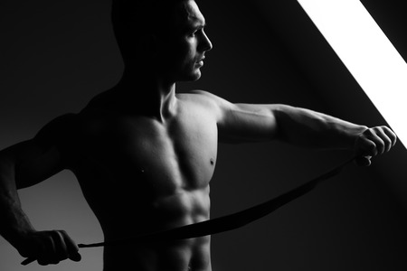 young man with muscular body and bare torso posing near window holding leather belt, black and whiteの写真素材