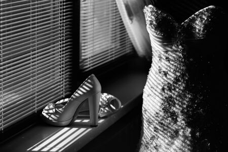 Beautiful elegant fashion bridal luxury wedding dress decorated beaded by ribbon hanging near window with blinds and shoes on heel standing near, black and whiteの写真素材