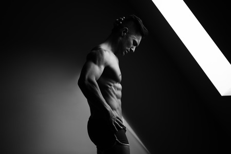 young man with muscular body and bare torso posing in shorts near window, black and whiteの写真素材