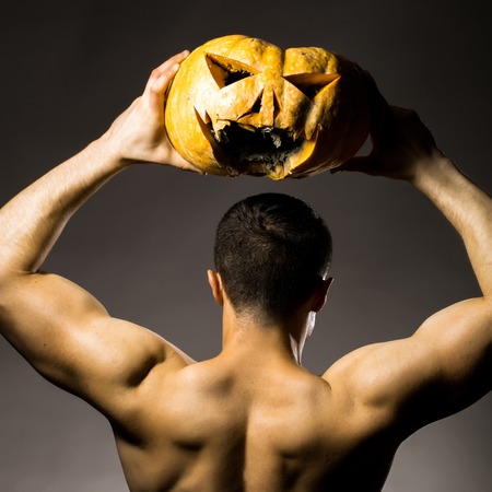 Young handsome man with muscular hands and back posing in studio holding yellow halloween pumpkin on grey backgroundの写真素材
