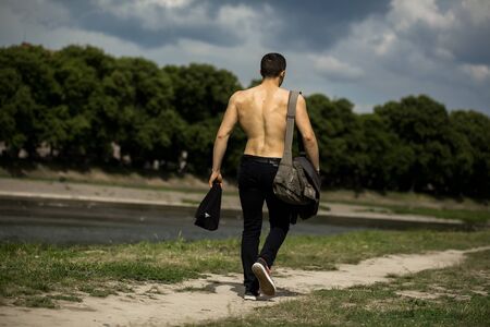 Young handsome man with muscular body and bare torso going on ground road near green grass with training bag outdoor with cloudy skyの写真素材