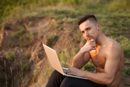 Young handsome man with muscular sexy body and bare chest sitting with laptop outdoor sunny dayの写真素材