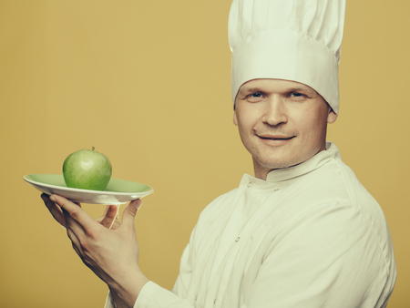 Young chef with green apple on plate in white uniform and cook hat on yellow backgroundの写真素材