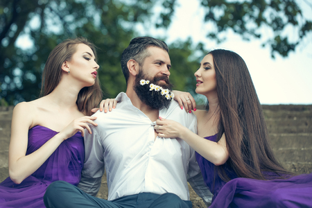handsome man in white shirt with dandelion flowers in beard with two young pretty women in violet dresses on stony stairs sunny day outdoorの写真素材