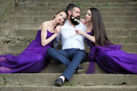 handsome man in white shirt with dandelion flowers in beard with two young pretty women in violet dresses on stony stairs sunny day outdoorの写真素材