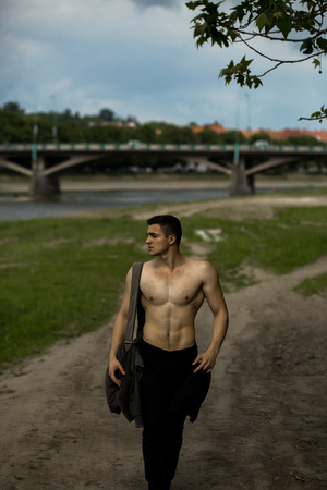 Young handsome man with muscular body and torso going on ground road near green grass with training bag on bare chest outdoorの写真素材